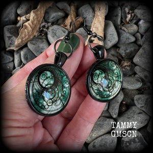 These awesome earrings feature a Dr Who inspired Gallifreyan steampunk sigil enclosed in a shiny resin dome, nice and dangly at just over 6cms from tip to tip, and weighing approx 8 grams a piece.
This pair has been made on french hooks, suitable for pierced ears.