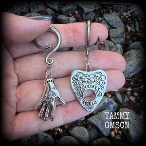 Featuring an antique silver Ouija planchette and esoteric hand charm, this pair of gorgeous earrings weigh approx 5 grams a piece, and measure 6cms from tip to tip.

This pair has been made on 6 gauge (4mm) surgical steel full curl hooks, suitable for stretched lobes.

