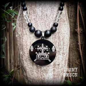 This necklace features gorgeous glass and matte black obsidian beads, with a large engraved enamel veve pendant, inspired by Baron Samedi.

This necklace measures approx 40cms when laying flat, and fastens with an antique silver toggle clasp.