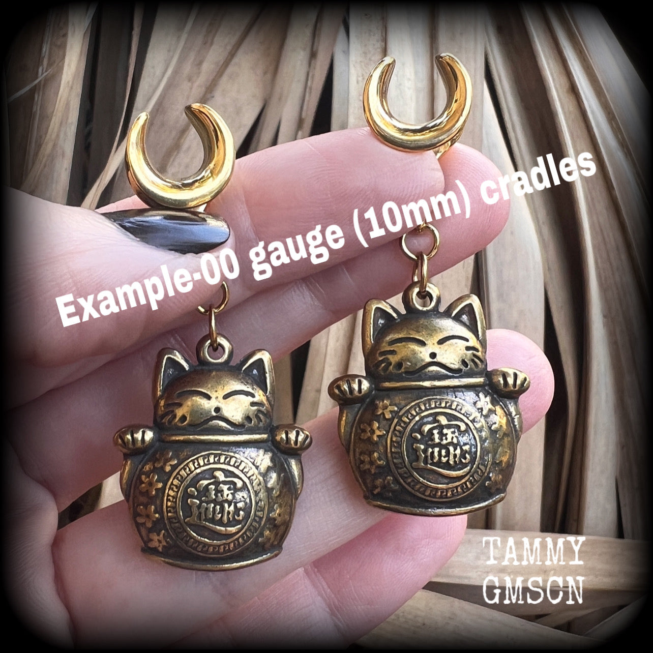 May include: Two gold-colored metal ear gauges with a dangling lucky cat charm. The charms are round and have a cat face with a coin in its paw. The gauges are a spiral shape and are hanging from a wooden surface.