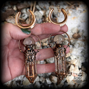 These unique ear weights feature big clear quartz points enclosed in a copper electroplate cage with topped with folk horror inspired celestial suns. These gorgeous earrings measure 11cms from tip to tip, and weigh approx 45 grams each.
This pair has been made on 1” gauge (25mm) rose gold tone surgical steel cradles, suitable for stretched lobes.