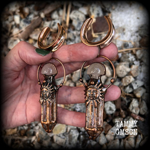 These unique ear weights feature big clear quartz points enclosed in a copper electroplate cage with topped with folk horror inspired celestial suns. These gorgeous earrings measure 11cms from tip to tip, and weigh approx 45 grams each.
This pair has been made on 1” gauge (25mm) rose gold tone surgical steel cradles, suitable for stretched lobes.