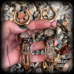 These unique ear weights feature big clear quartz points enclosed in a copper electroplate cage with topped with folk horror inspired celestial suns. These gorgeous earrings measure 11cms from tip to tip, and weigh approx 45 grams each.
This pair has been made on 1” gauge (25mm) rose gold tone surgical steel cradles, suitable for stretched lobes.