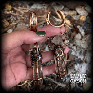 These unique ear weights feature big clear quartz points enclosed in a copper electroplate cage with topped with folk horror inspired celestial suns. These gorgeous earrings measure 11cms from tip to tip, and weigh approx 45 grams each.
This pair has been made on 1” gauge (25mm) rose gold tone surgical steel cradles, suitable for stretched lobes.