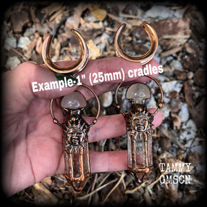 These unique ear weights feature big clear quartz points enclosed in a copper electroplate cage with topped with folk horror inspired celestial suns. These gorgeous earrings measure 11cms from tip to tip, and weigh approx 45 grams each.
This pair has been made on 1” gauge (25mm) rose gold tone surgical steel cradles, suitable for stretched lobes.