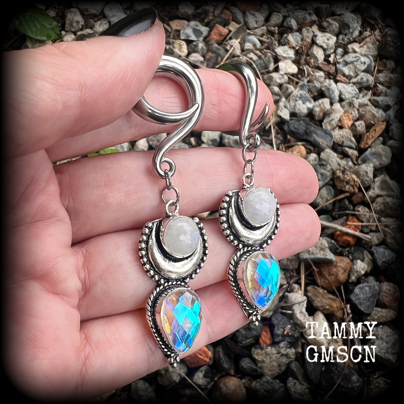 May include: Two silver ear gauges with dangling charms. Each charm has a rainbow moonstone, a silver crescent moon, and a teardrop-shaped iridescent gemstone. These earrings are available on a selection of different sized hooks.