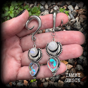 May include: Two silver ear gauges with dangling charms. Each charm has a rainbow moonstone, a silver crescent moon, and a teardrop-shaped iridescent gemstone. These earrings are available on a selection of different sized hooks.