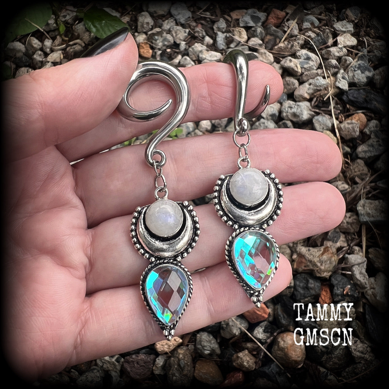 May include: Two silver ear gauges with dangling charms. Each charm has a rainbow moonstone, a silver crescent moon, and a teardrop-shaped iridescent gemstone. These earrings are available on a selection of different sized hooks.