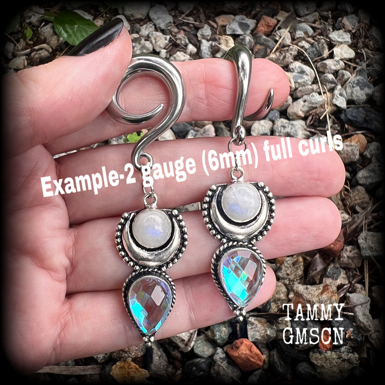 May include: Two silver ear gauges with dangling charms. Each charm has a rainbow moonstone, a silver crescent moon, and a teardrop-shaped iridescent gemstone. These earrings are available on a selection of different sized hooks.