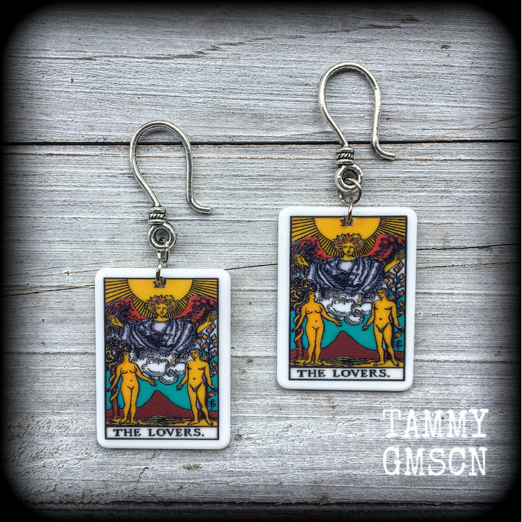 May include: Two silver earrings with a white rectangular pendant featuring a tarot card image of The Lovers. The card features a yellow sun, a winged figure with a crown, and two nude figures standing on either side of a mountain with a red peak. The text on the card reads 'THE LOVERS.'