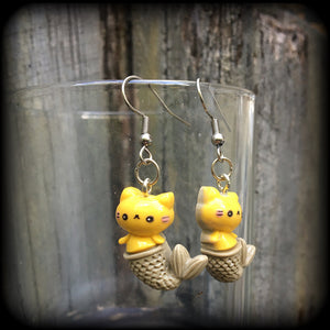 May include: A pair of earrings with a yellow cat mermaid design. The earrings are made of acrylic and have a silver hook. The cat mermaid has a smiley face, and a brown tail with scales. 