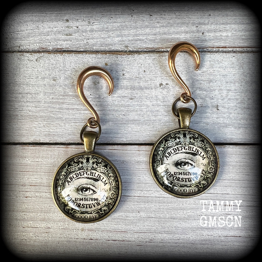 These gorgeous one of a kind Ouija board gauged earrings feature an antique bronze cabachon charm showing a classic ouija board image complete with the all seeing Mystic eye.

These earrings measure just on 6.5cms from tip to tip, and weigh approx 11 grams each.

This pair has been made on 6 gauge (4mm) stainless steel half curl hooks, suitable for stretched lobes.