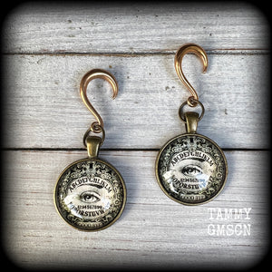 These gorgeous one of a kind Ouija board gauged earrings feature an antique bronze cabachon charm showing a classic ouija board image complete with the all seeing Mystic eye.

These earrings measure just on 6.5cms from tip to tip, and weigh approx 11 grams each.

This pair has been made on 6 gauge (4mm) stainless steel half curl hooks, suitable for stretched lobes.
