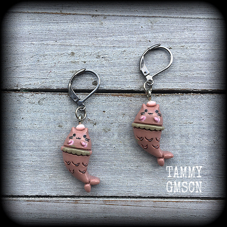 May include: A pair of earrings with a pink cat mermaid design. The earrings are made of acrylic and have a silver leverback hook. The cat mermaid has a pink face, and a brown tail with scales. 