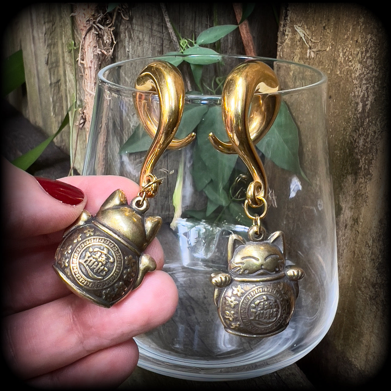 May include: Two gold-colored metal ear gauges with a dangling lucky cat charm. The charms are round and have a cat face with a coin in its paw. The gauges are a spiral shape and are hanging from a wooden surface.