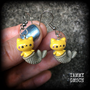 May include: A pair of earrings with a yellow cat mermaid design. The earrings are made of acrylic and have a silver hook. The cat mermaid has a smiley face, and a brown tail with scales. 