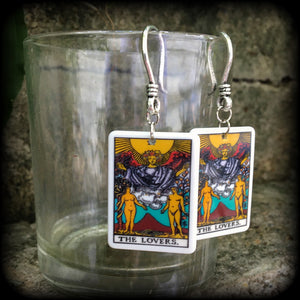 May include: Two silver earrings with a white rectangular pendant featuring a tarot card image of The Lovers. The card features a yellow sun, a winged figure with a crown, and two nude figures standing on either side of a mountain with a red peak. The text on the card reads 'THE LOVERS.'