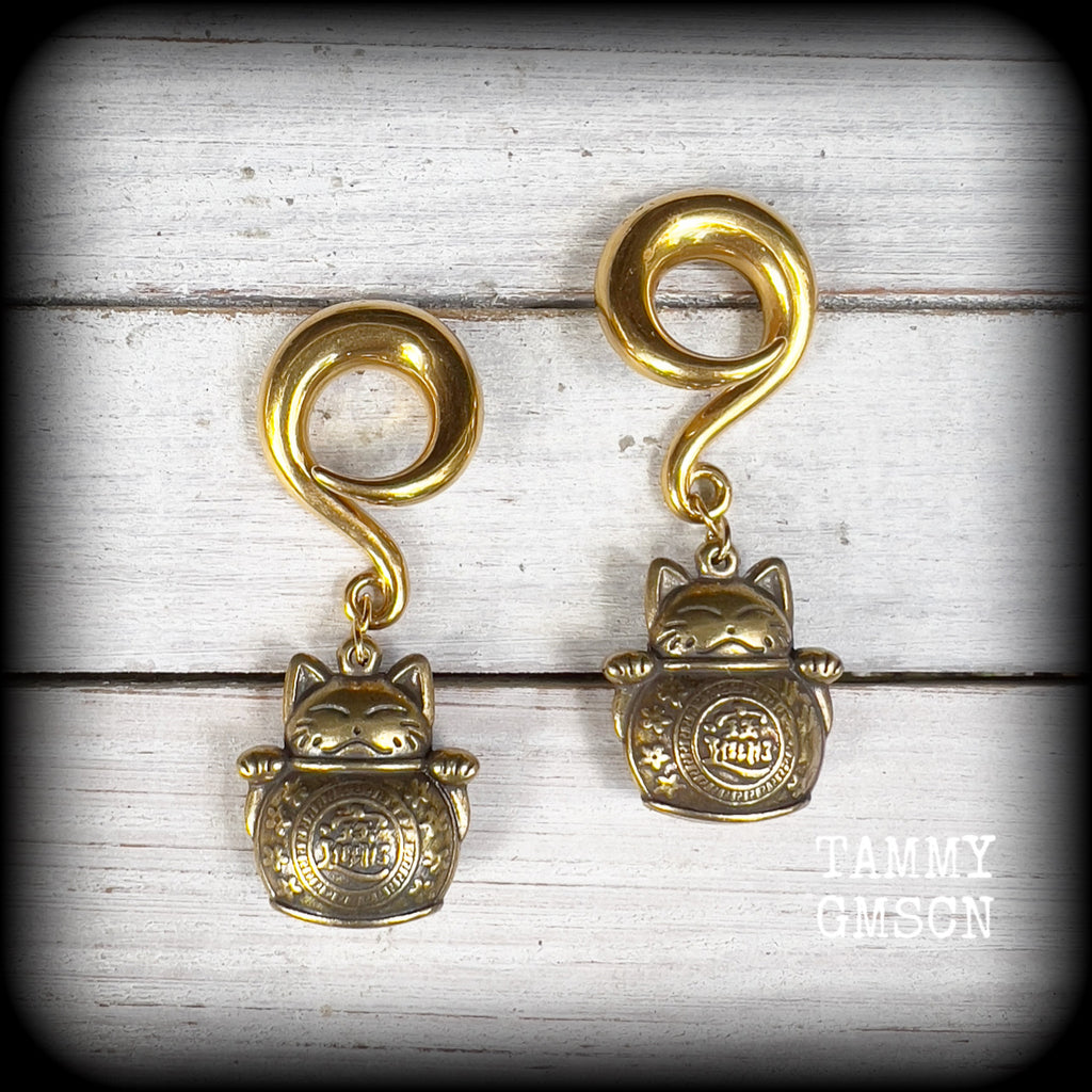 May include: Two gold-colored metal ear gauges with a dangling lucky cat charm. The charms are round and have a cat face with a coin in its paw. The gauges are a spiral shape and are hanging from a wooden surface.