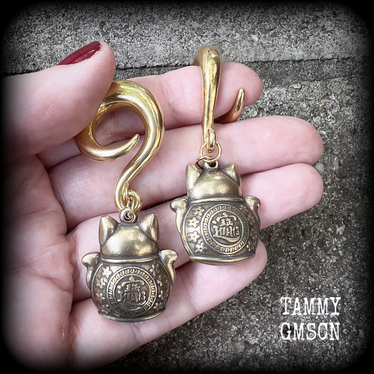 May include: Two gold-colored metal ear gauges with a dangling lucky cat charm. The charms are round and have a cat face with a coin in its paw. The gauges are a spiral shape and are hanging from a wooden surface.