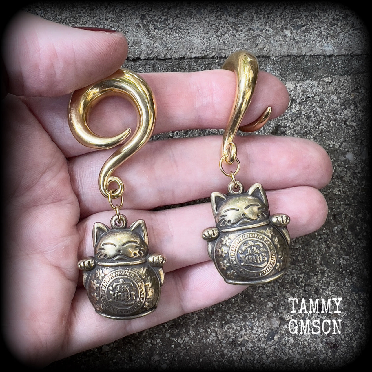 May include: Two gold-colored metal ear gauges with a dangling lucky cat charm. The charms are round and have a cat face with a coin in its paw. The gauges are a spiral shape and are hanging from a wooden surface.