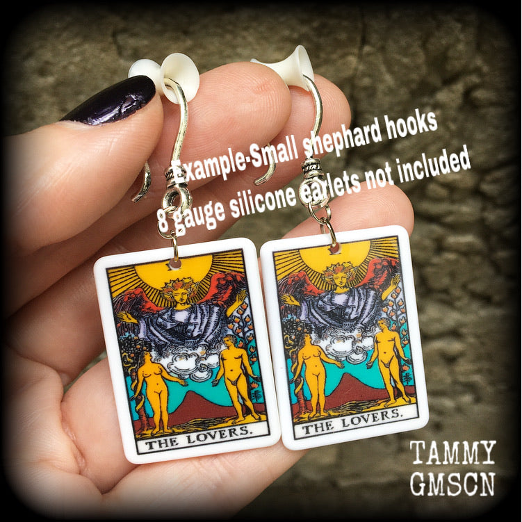 May include: Two silver earrings with a white rectangular pendant featuring a tarot card image of The Lovers. The card features a yellow sun, a winged figure with a crown, and two nude figures standing on either side of a mountain with a red peak. The text on the card reads 'THE LOVERS.'