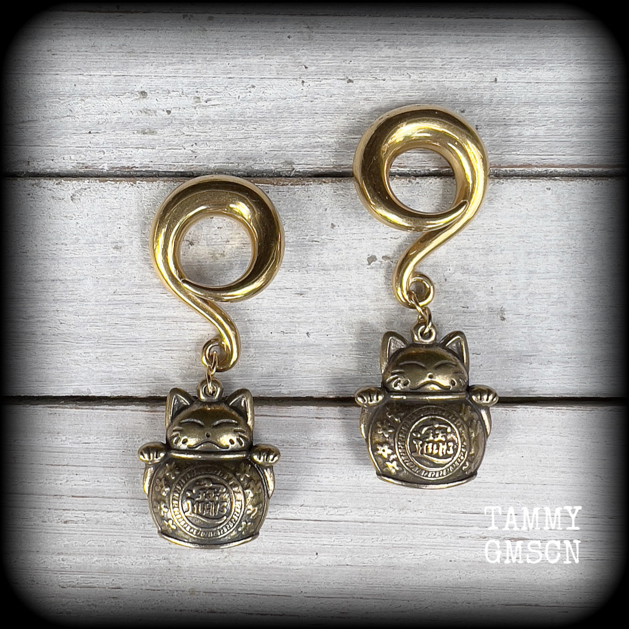 May include: Two gold-colored metal ear gauges with a dangling lucky cat charm. The charms are round and have a cat face with a coin in its paw. The gauges are a spiral shape and are hanging from a wooden surface.