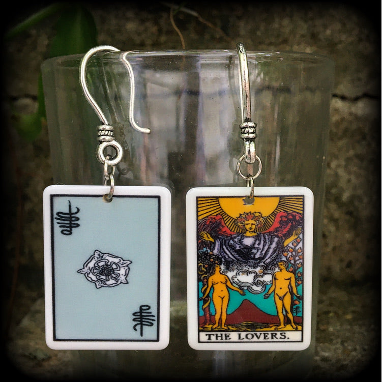 May include: Two silver earrings with a white rectangular pendant featuring a tarot card image of The Lovers. The card features a yellow sun, a winged figure with a crown, and two nude figures standing on either side of a mountain with a red peak. The text on the card reads 'THE LOVERS.'