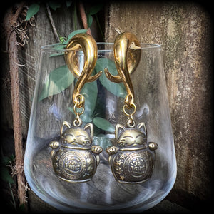 May include: Two gold-colored metal ear gauges with a dangling lucky cat charm. The charms are round and have a cat face with a coin in its paw. The gauges are a spiral shape and are hanging from a wooden surface.