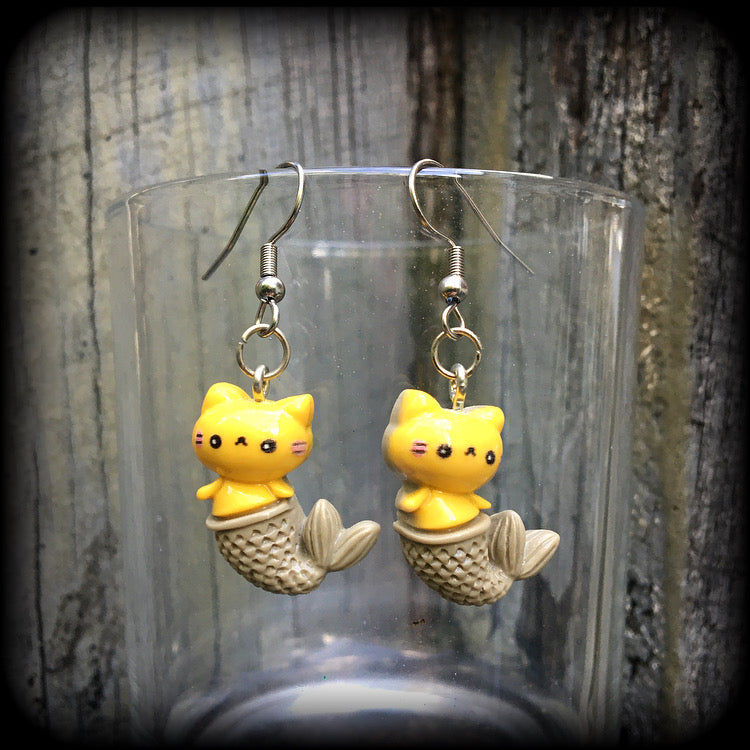 May include: A pair of earrings with a yellow cat mermaid design. The earrings are made of acrylic and have a silver hook. The cat mermaid has a smiley face, and a brown tail with scales. 