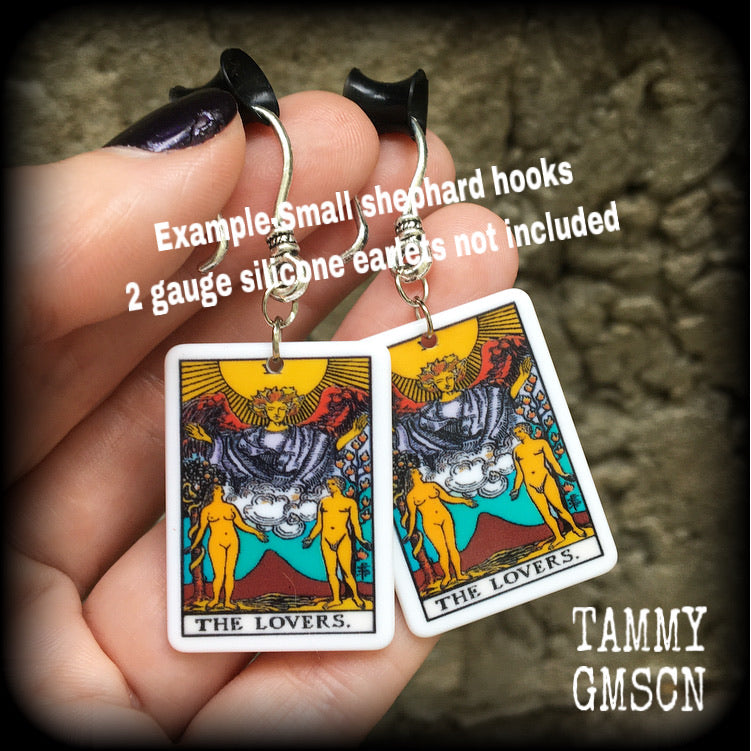 May include: Two silver earrings with a white rectangular pendant featuring a tarot card image of The Lovers. The card features a yellow sun, a winged figure with a crown, and two nude figures standing on either side of a mountain with a red peak. The text on the card reads 'THE LOVERS.'