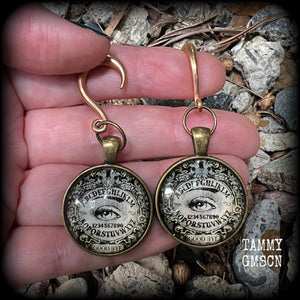 These gorgeous one of a kind Ouija board gauged earrings feature an antique bronze cabachon charm showing a classic ouija board image complete with the all seeing Mystic eye.

These earrings measure just on 6.5cms from tip to tip, and weigh approx 11 grams each.

This pair has been made on 6 gauge (4mm) stainless steel half curl hooks, suitable for stretched lobes.