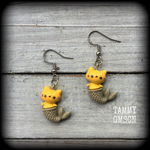 May include: A pair of earrings with a yellow cat mermaid design. The earrings are made of acrylic and have a silver hook. The cat mermaid has a smiley face, and a brown tail with scales. 