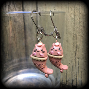 May include: A pair of earrings with a pink cat mermaid design. The earrings are made of acrylic and have a silver leverback hook. The cat mermaid has a pink face, and a brown tail with scales. 