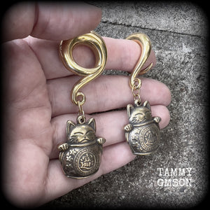 May include: Two gold-colored metal ear gauges with a dangling lucky cat charm. The charms are round and have a cat face with a coin in its paw. The gauges are a spiral shape and are hanging from a wooden surface.