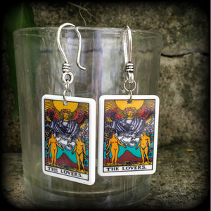 May include: Two silver earrings with a white rectangular pendant featuring a tarot card image of The Lovers. The card features a yellow sun, a winged figure with a crown, and two nude figures standing on either side of a mountain with a red peak. The text on the card reads 'THE LOVERS.'