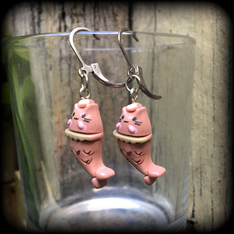 May include: A pair of earrings with a pink cat mermaid design. The earrings are made of acrylic and have a silver leverback hook. The cat mermaid has a pink face, and a brown tail with scales. 