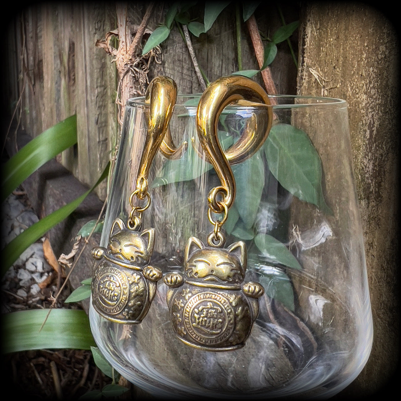 May include: Two gold-colored metal ear gauges with a dangling lucky cat charm. The charms are round and have a cat face with a coin in its paw. The gauges are a spiral shape and are hanging from a wooden surface.