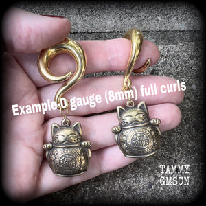 May include: Two gold-colored metal ear gauges with a dangling lucky cat charm. The charms are round and have a cat face with a coin in its paw. The gauges are a spiral shape and are hanging from a wooden surface.