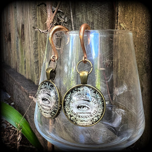 These gorgeous one of a kind Ouija board gauged earrings feature an antique bronze cabachon charm showing a classic ouija board image complete with the all seeing Mystic eye.

These earrings measure just on 6.5cms from tip to tip, and weigh approx 11 grams each.

This pair has been made on 6 gauge (4mm) stainless steel half curl hooks, suitable for stretched lobes.